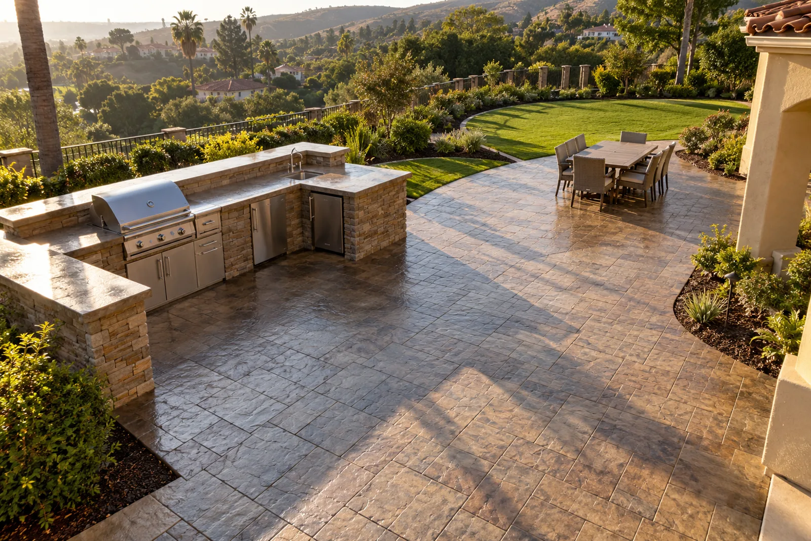 Stamped slate-pattern concrete patio in warm sandstone tones with a built-in outdoor kitchen island and dining table on a hillside lot