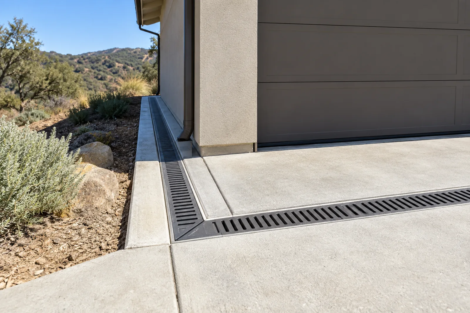 Black-grated trench drain across a concrete driveway at a garage, connected to a concrete swale along the side of the house