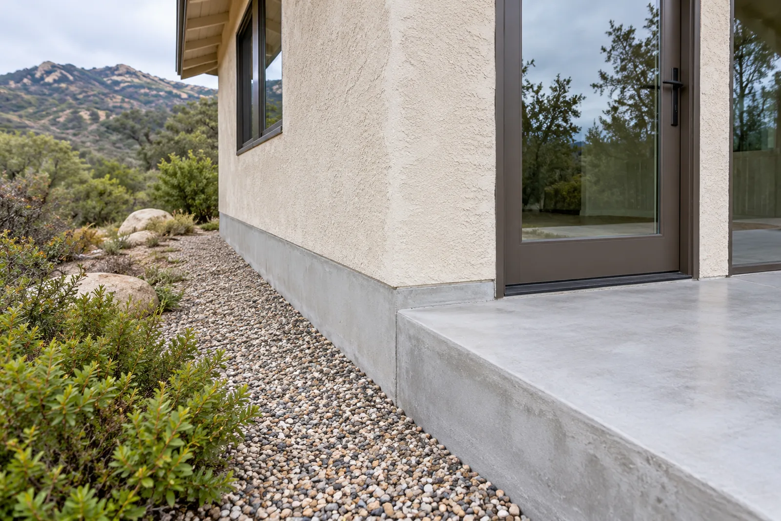 Clean intact concrete stem wall and slab edge at the corner of a stucco house beside a gravel strip, with chaparral hills behind