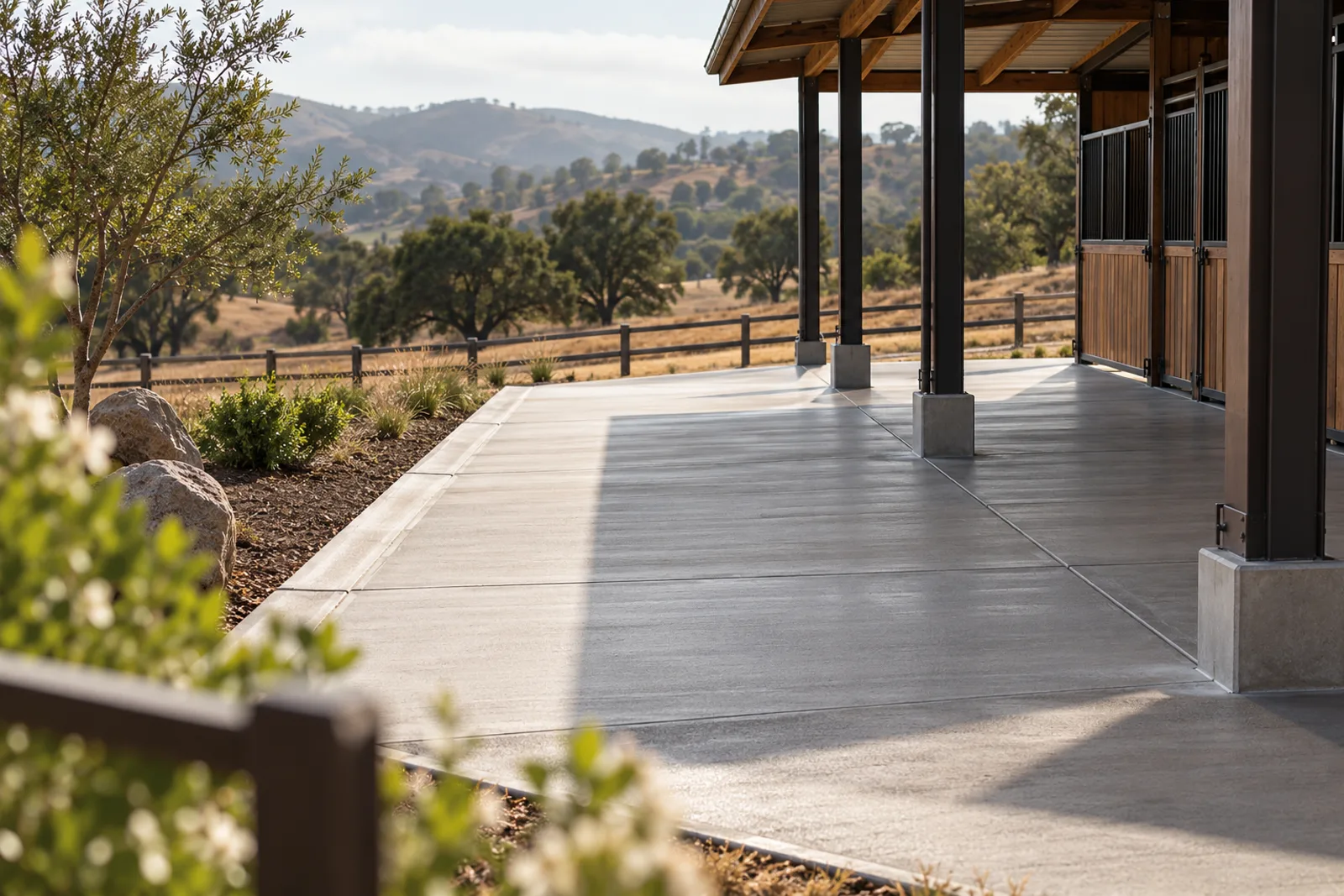Broom-finished concrete aisle and wash area under a barn roof with thickened edges at the posts, overlooking rolling pasture